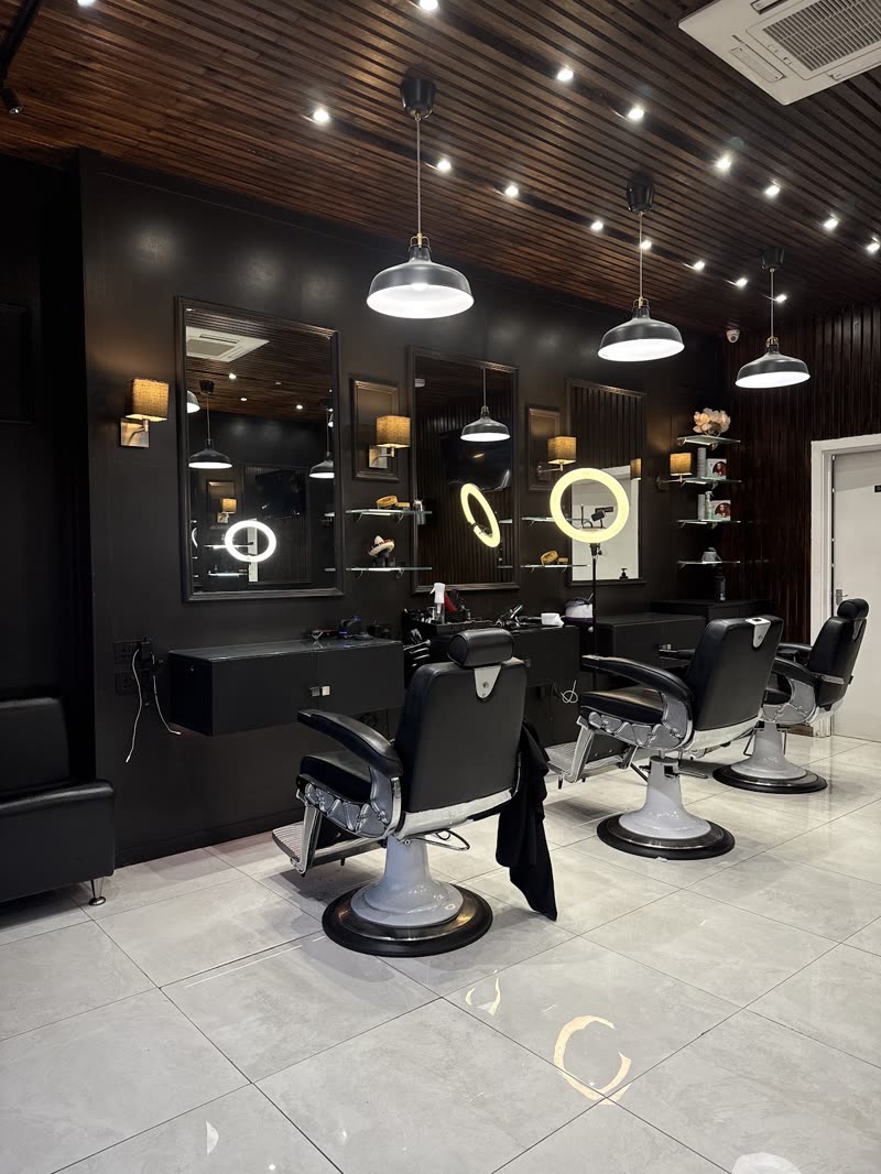 The salon interior: three black leather barber chairs on chrome bases in front of backlit round mirrors, under a dark slatted ceiling