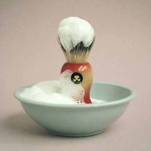 A shaving brush standing in a bowl of white lather on a plain pastel surface